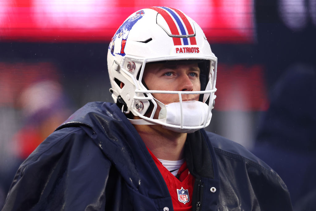 A football player in a Patriots helmet and a blue jacket looks upward. The helmet features a blue and red stripe and a detailed logo on the side.