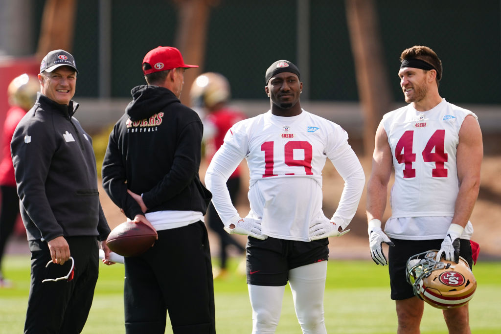 Four men are standing on a football field. Two are wearing 49ers jerseys, one holds a helmet, another a football. They're talking and smiling casually.