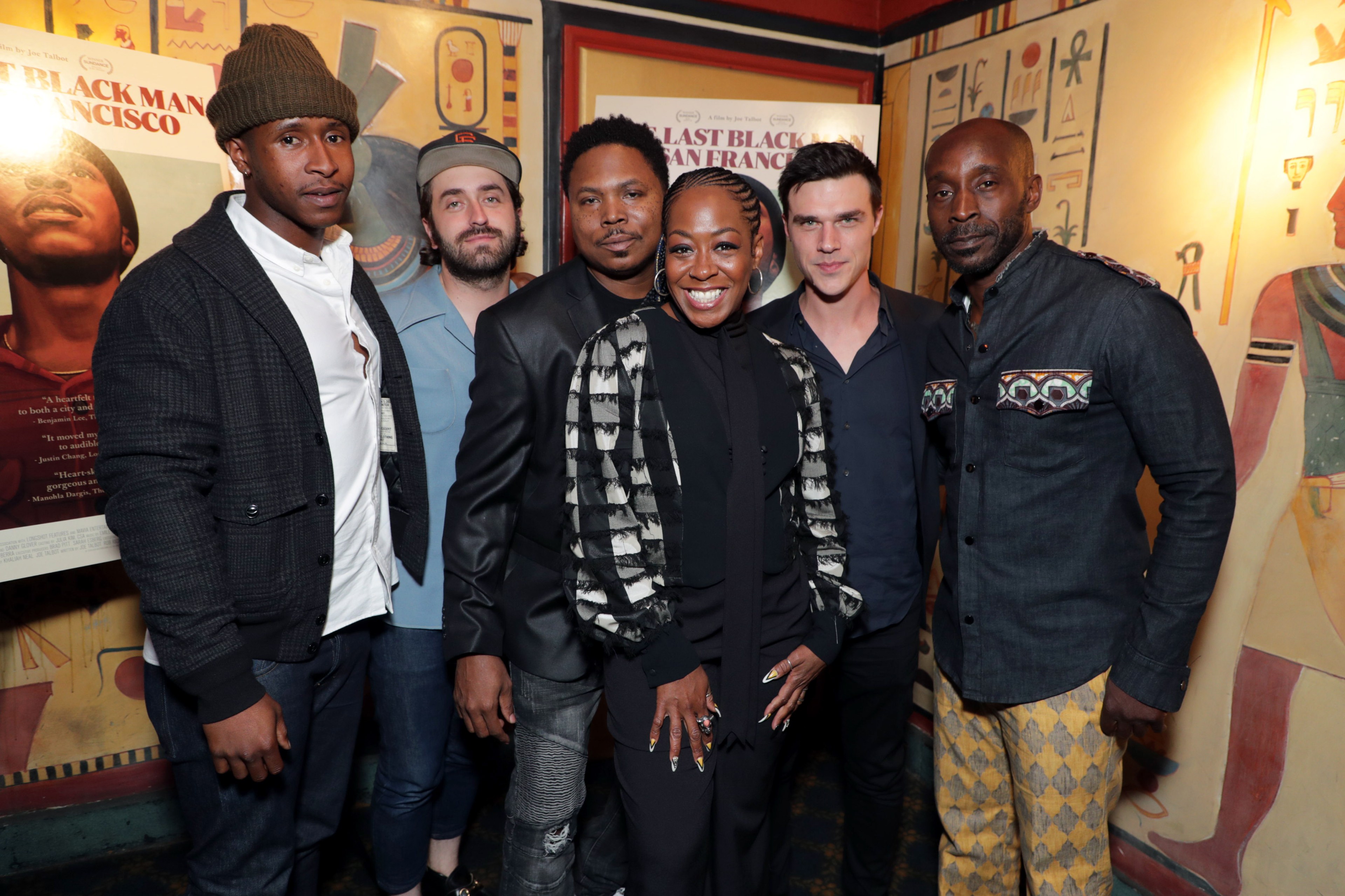 Six people stand smiling in front of wall art and a movie poster. They are dressed in stylish outfits, and the atmosphere appears celebratory.