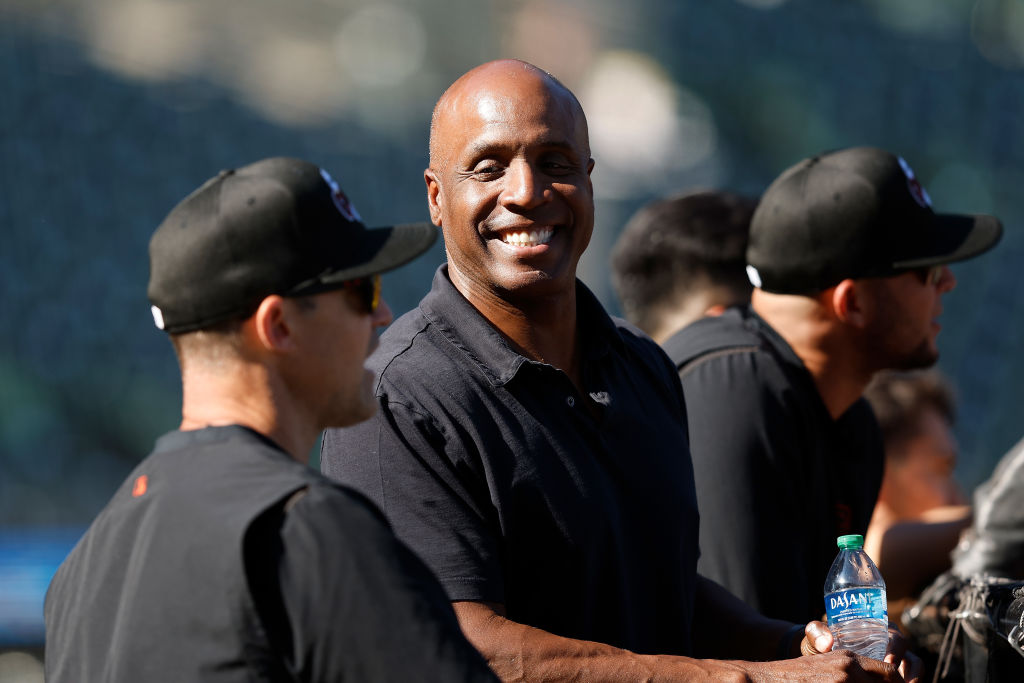 A man in a black shirt smiles brightly while holding a water bottle. He's surrounded by others in caps, possibly at a sports event.