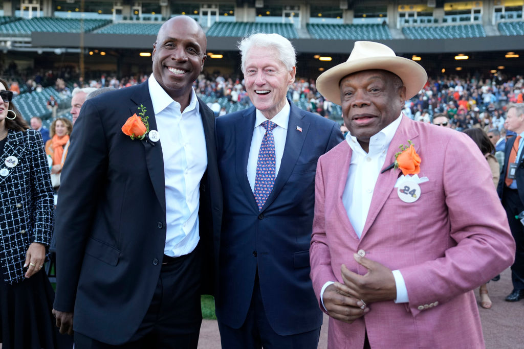 Three men are standing together, smiling. The two on the sides wear suits with orange flowers, and the one in the middle has a blue suit and red tie. A crowd is behind them.