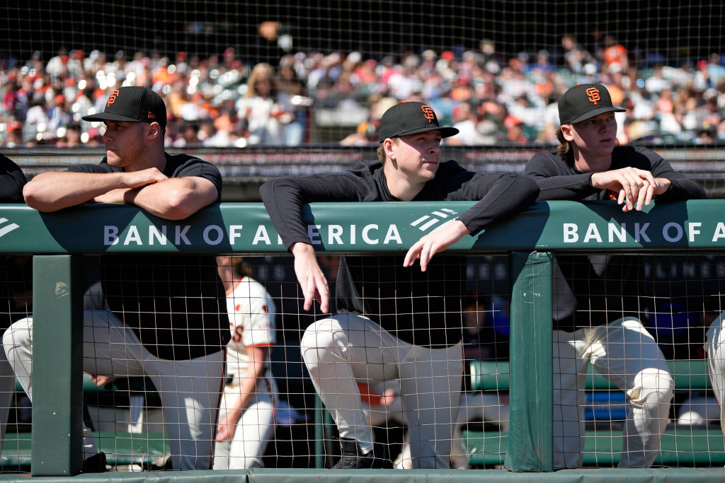 Three baseball players in black jerseys and caps lean on a dugout fence with "Bank of America" signage, watching a game as a crowd sits in the background.