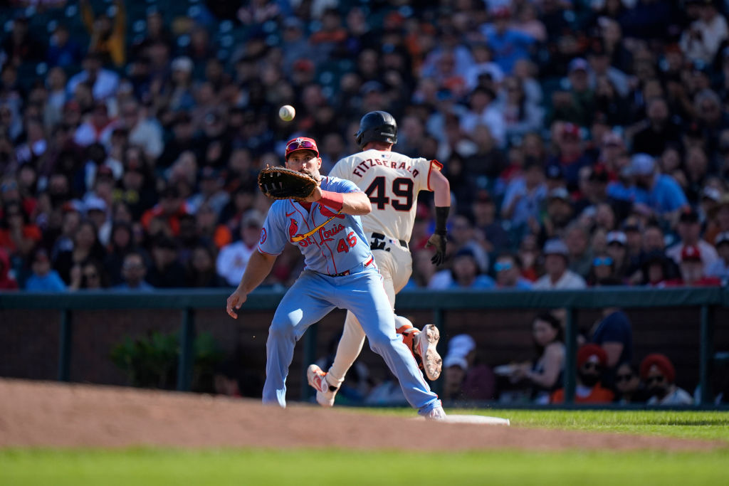 A baseball player in a red uniform attempts to catch a ball near first base as another player in a white uniform runs past. A crowd watches intently.