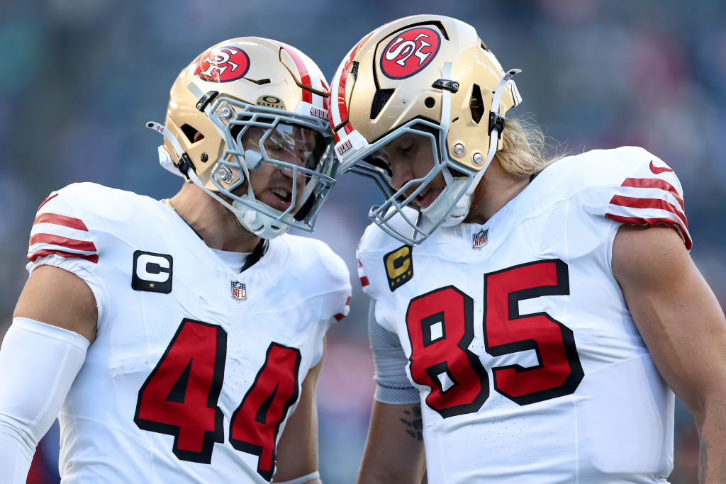 Two football players in white 49ers uniforms with red numbers and gold helmets are touching helmets, appearing focused and intense.