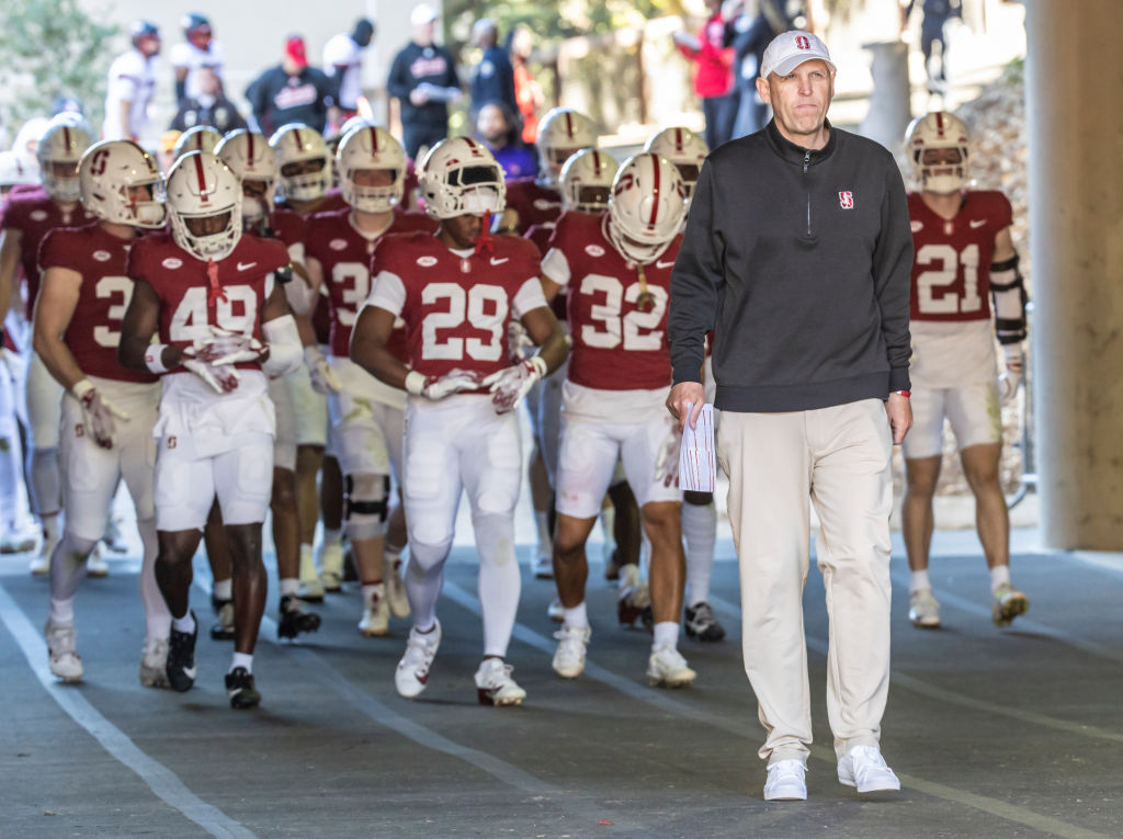 A football coach leads a team wearing red jerseys and white pants out of a tunnel. The players have helmets on, and the scene is bright with a focused intensity.