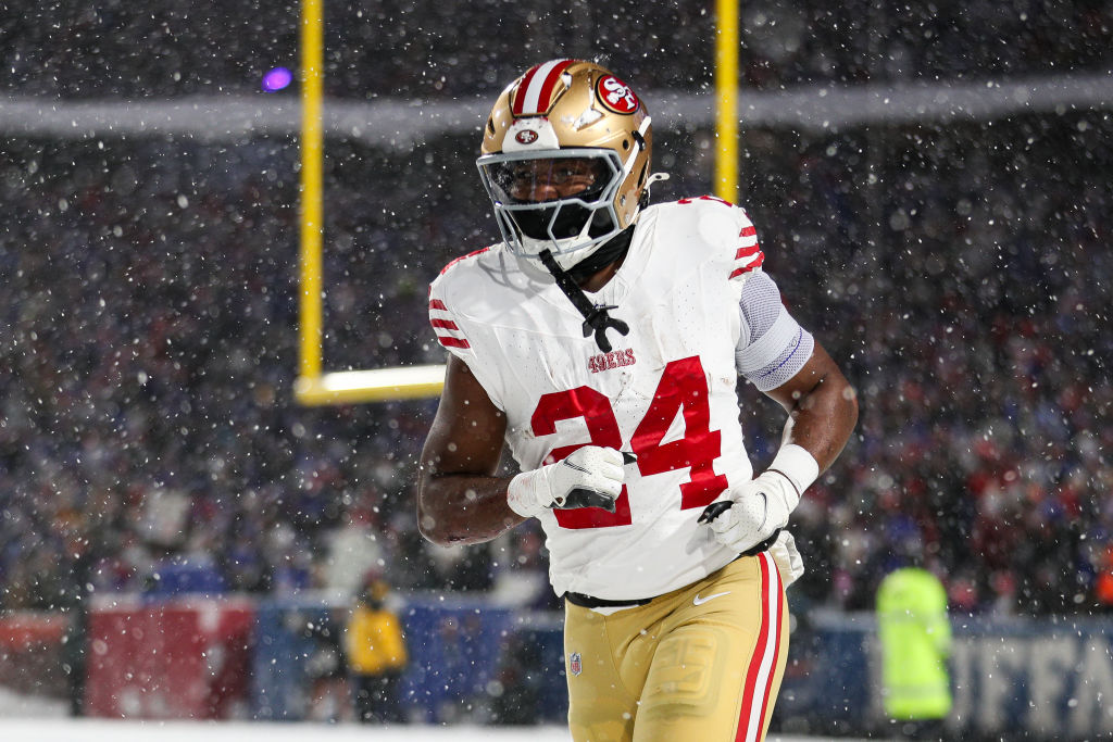 A football player in a white and red 49ers uniform runs on a snowy field. The background is blurred, showing an audience and goalposts under snowfall.