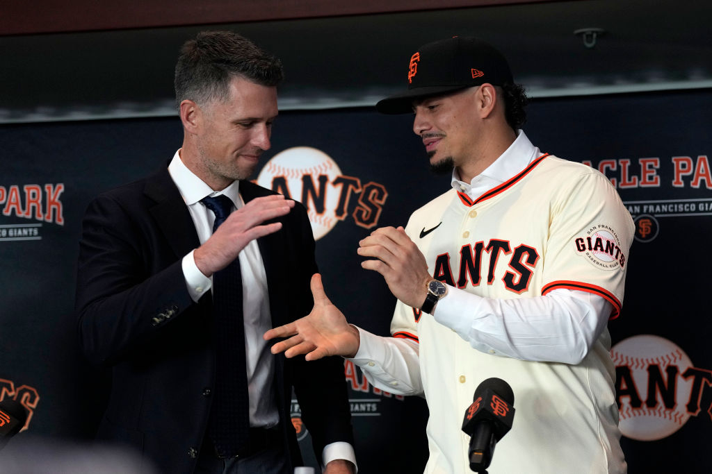 Two men shake hands in front of a &quot;San Francisco Giants&quot; backdrop. One wears a suit, the other a Giants jersey and cap. They seem to be at a press event.