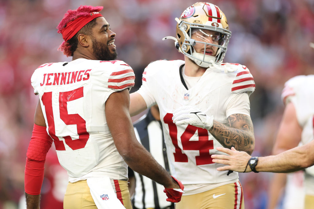 Two football players in 49ers uniforms are smiling on the field. Number 15 wears a red headband, while number 14 has tattoos and a helmet with a visor.