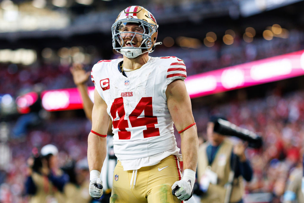 A football player in a white jersey with number 44 is cheering on the field. He's wearing a helmet, has his fists clenched, and the background is a blurred stadium.