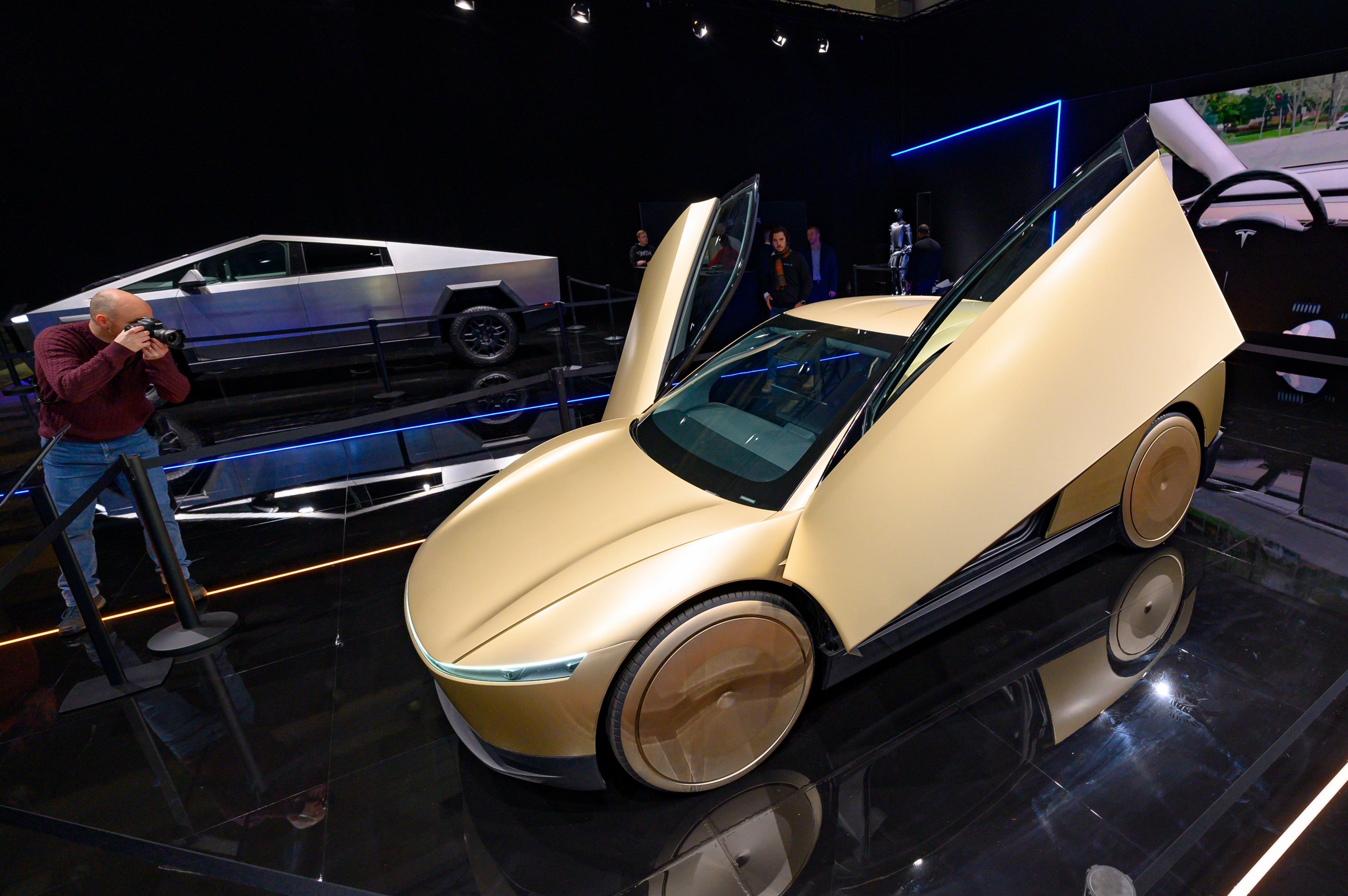 A futuristic gold car with open doors is displayed on a shiny black floor, with a person taking photos. A futuristic silver vehicle is visible in the background.