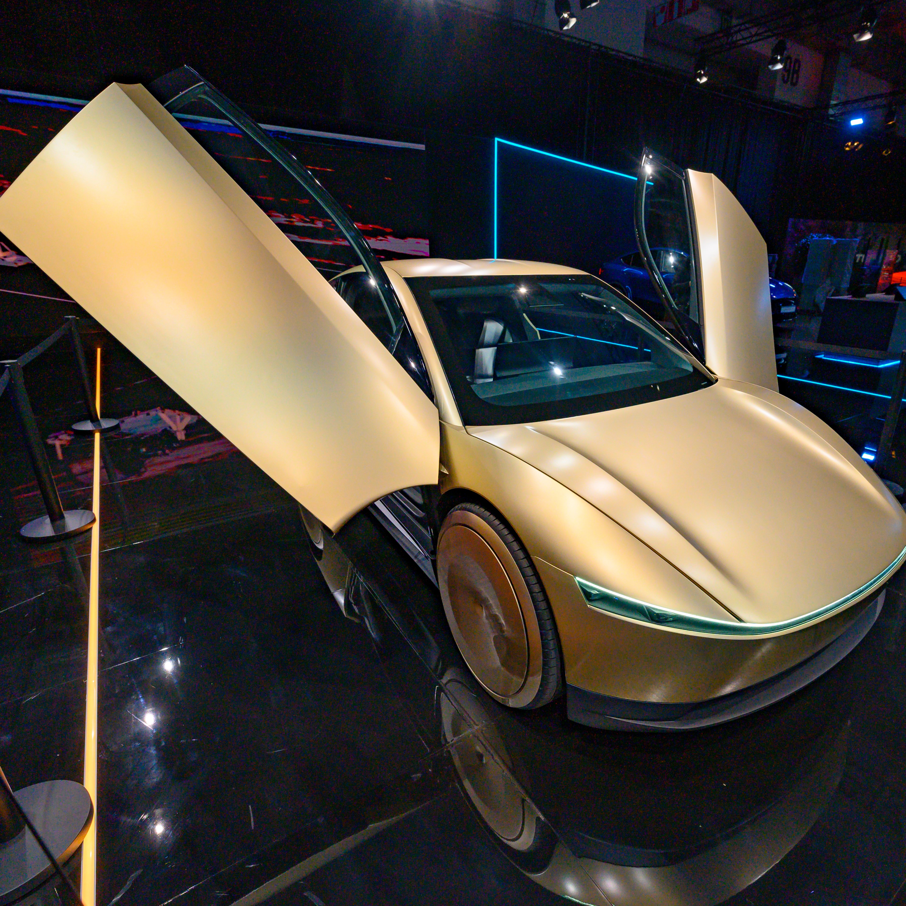 A sleek, gold-colored concept car with gullwing doors open, displayed on a shiny black reflective floor, surrounded by a minimalist exhibition setup.