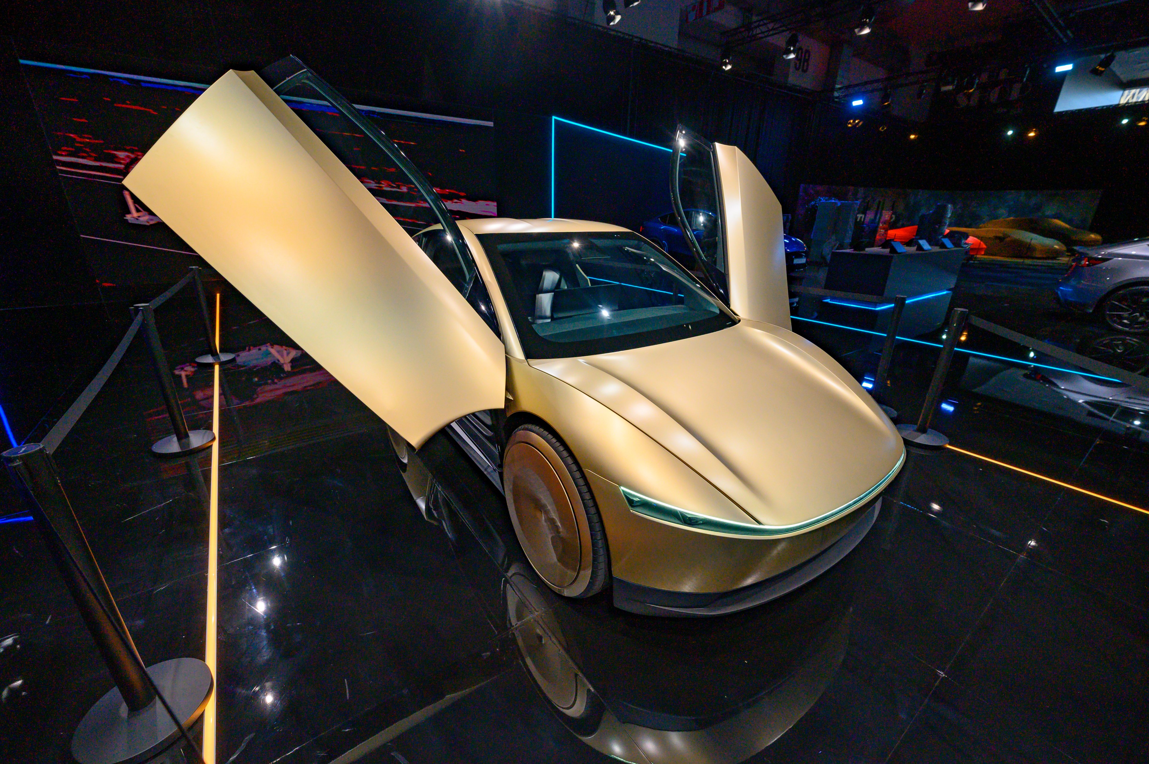 A sleek, gold-colored concept car with gullwing doors open, displayed on a shiny black reflective floor, surrounded by a minimalist exhibition setup.