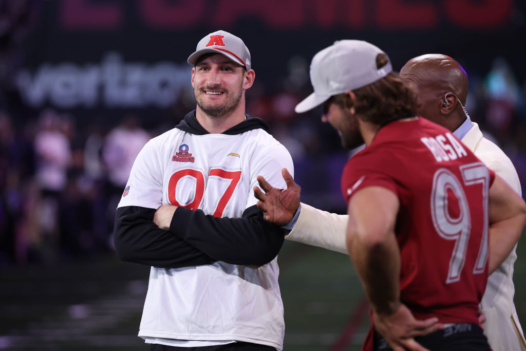 Three men are visible. One in the foreground wears a red shirt with "97," another in a white shirt also marked "97" smiles, and a third man in a suit is speaking.