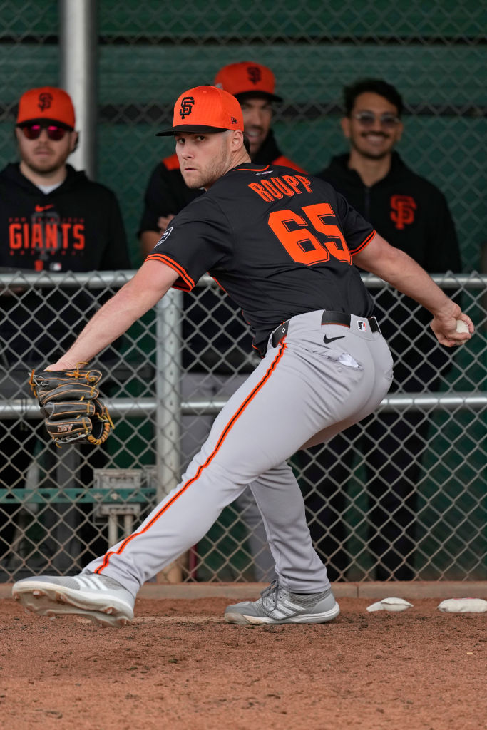 A baseball player is pitching on a mound, wearing a black and orange uniform. Several teammates in matching attire watch from behind a chain-link fence.