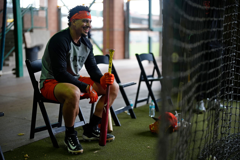 A person in sports attire sits on a folding chair with a baseball bat. They're wearing red gloves and an orange headband, smiling in a casual setting.