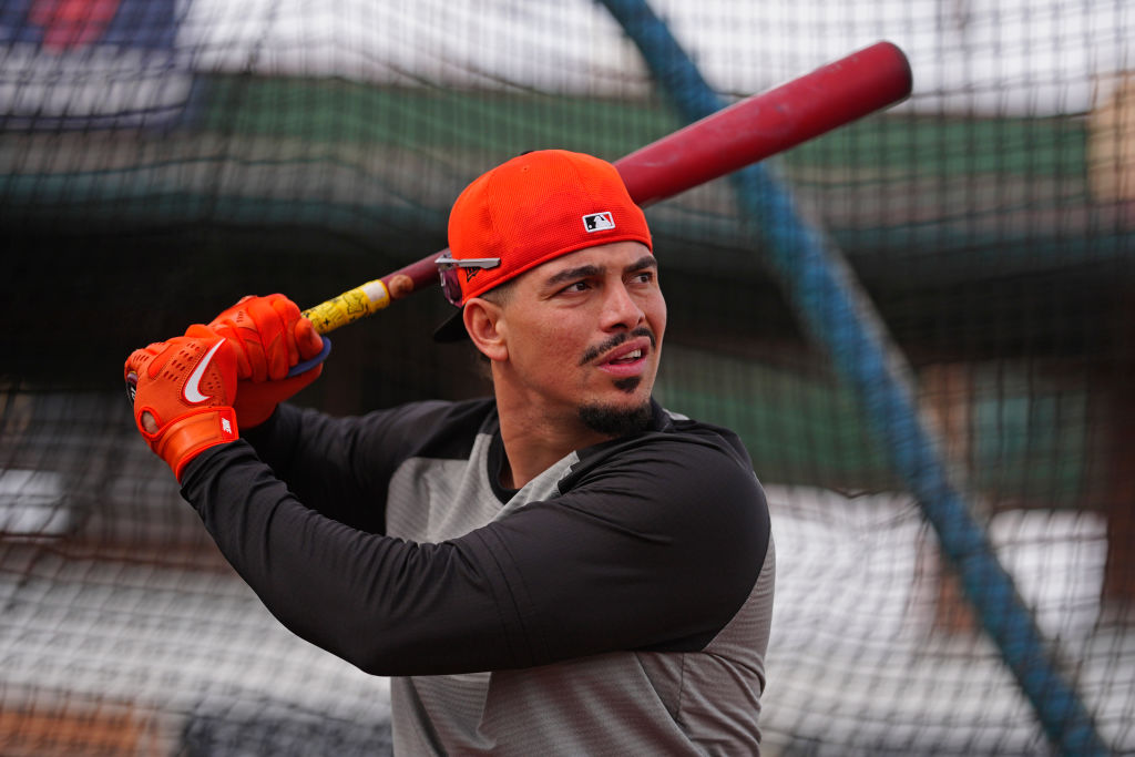 A person wearing an orange cap and gloves is holding a baseball bat, preparing to swing. They are in a batting cage with a focused expression.