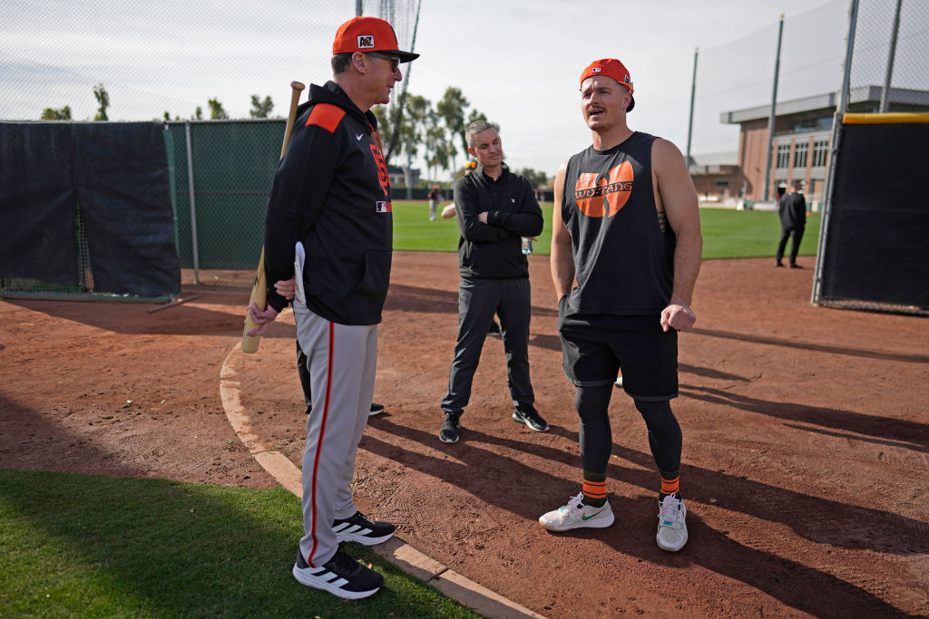 Several people stand on a baseball field. One person in athletic gear is talking animatedly to two others dressed in sports attire. A baseball bat leans on one person.
