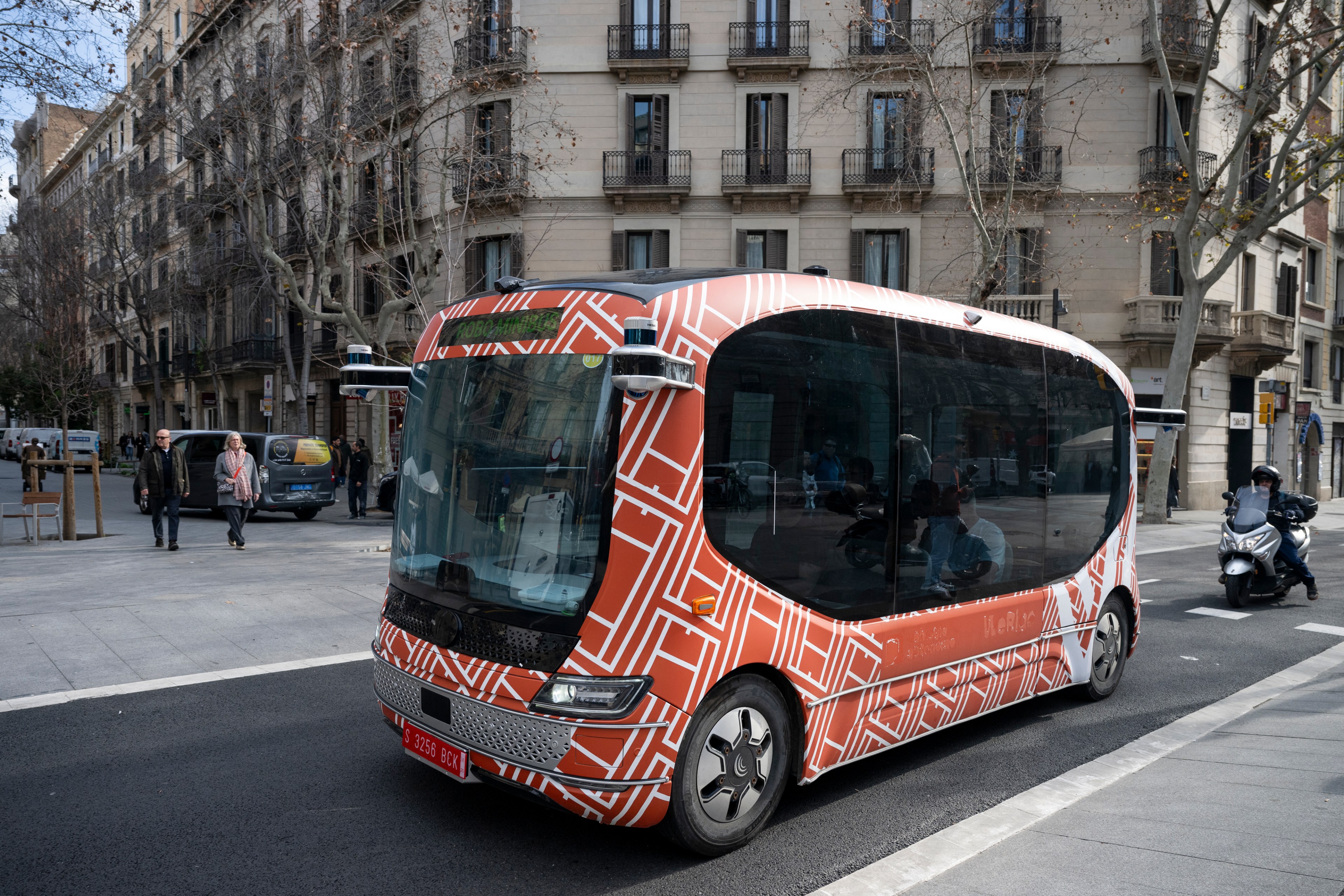 The image shows an autonomous bus with a geometric pattern design on an urban street. It's surrounded by pedestrians and other vehicles in a city area.