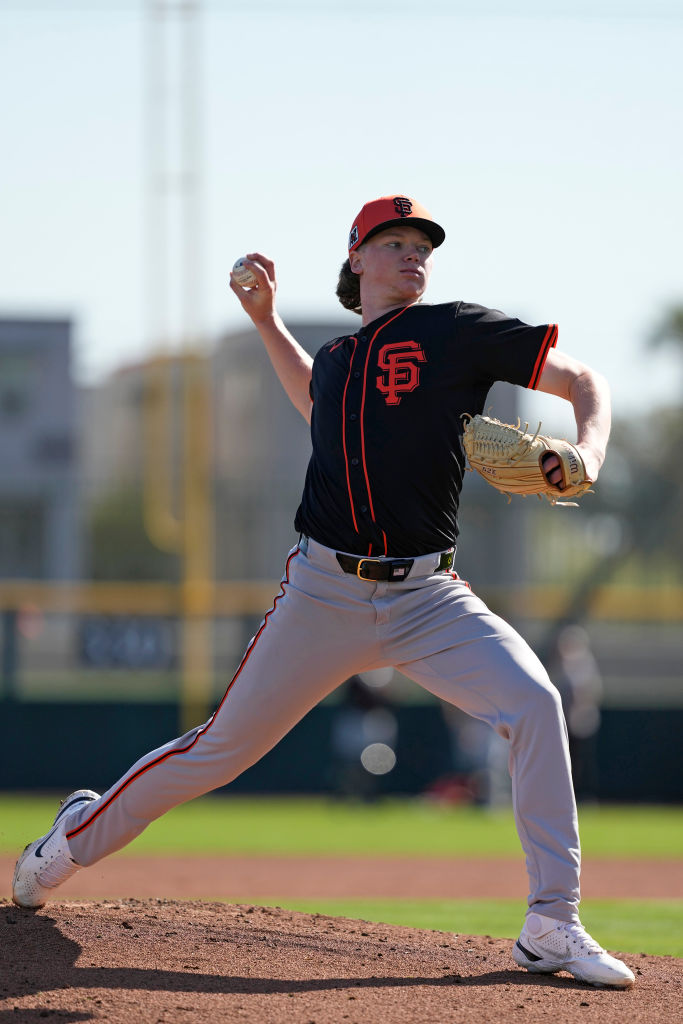 A baseball player in a black jersey and red cap is pitching on the mound, mid-throw, with a blurred background of a field and trees.