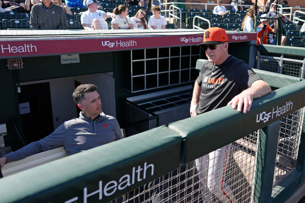 Two men are in a baseball dugout; one is seated wearing a zip-up jacket, and the other stands with sunglasses and a cap. Spectators sit in the stands behind them.