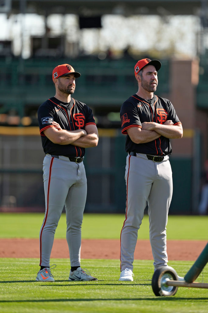 Two men in black and orange baseball uniforms stand on a field with arms crossed. They wear matching caps and are next to a baseball practice net on wheels.