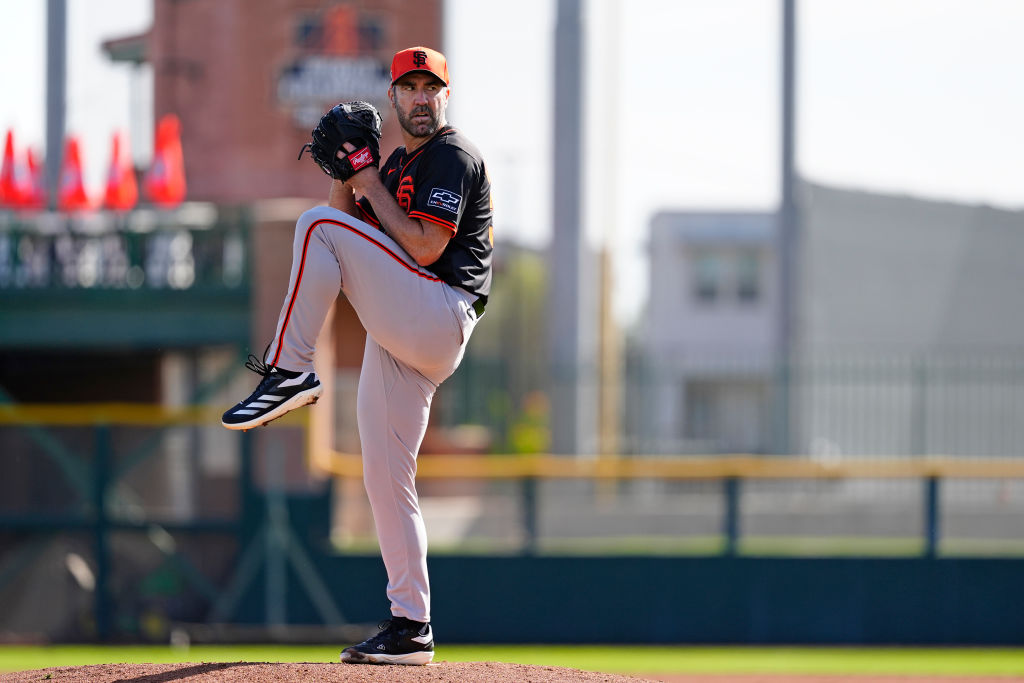 A baseball pitcher in a black and orange uniform stands on the mound, poised to throw. The background shows a blurred outfield fence and buildings.