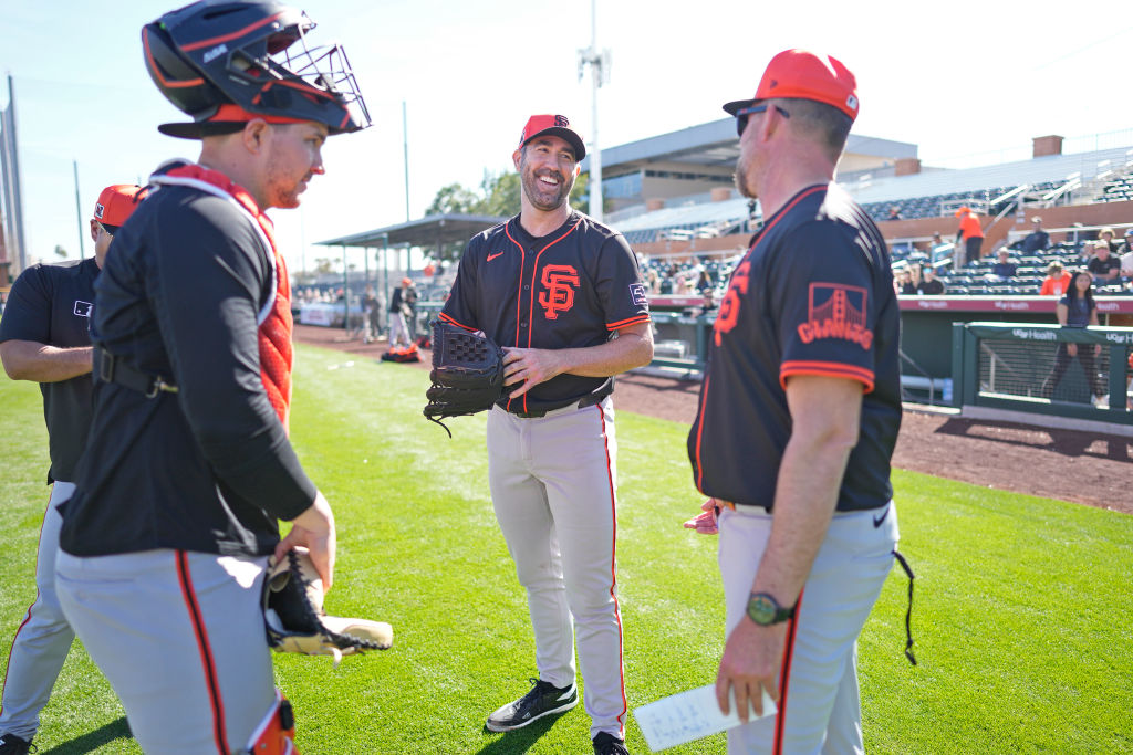 Three baseball players in black and orange uniforms stand on a field, talking and smiling. One wears catcher's gear. The sunny stadium is in the background.