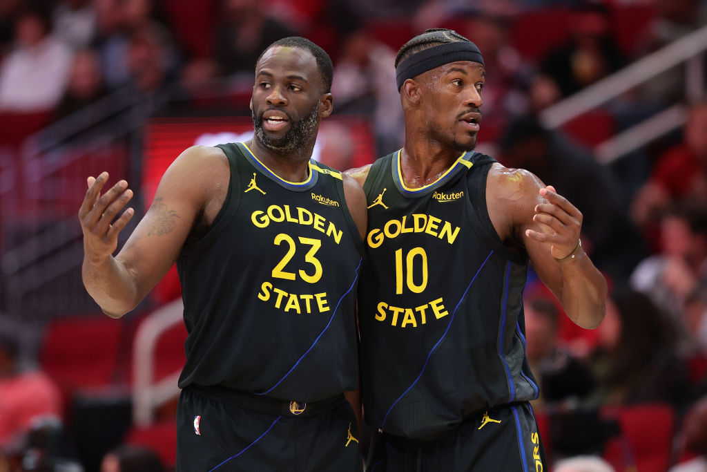 Two basketball players in dark jerseys with &quot;Golden State&quot; are on the court. One wears number 23, and the other 10, both gesturing and focused on the game.