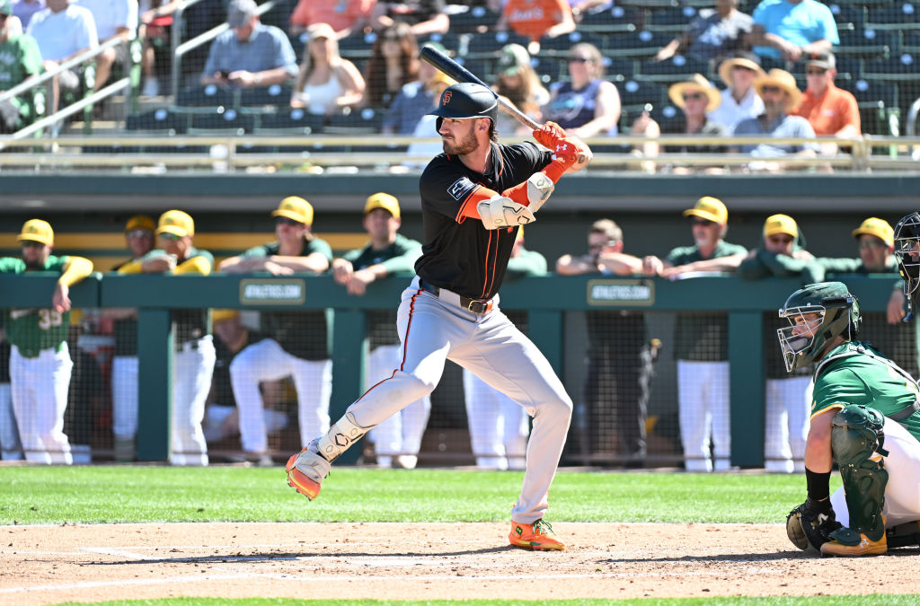 A baseball player in a black jersey is mid-swing at the plate. Nearby, a catcher in a green uniform crouches. Fans watch from bleachers in the background.