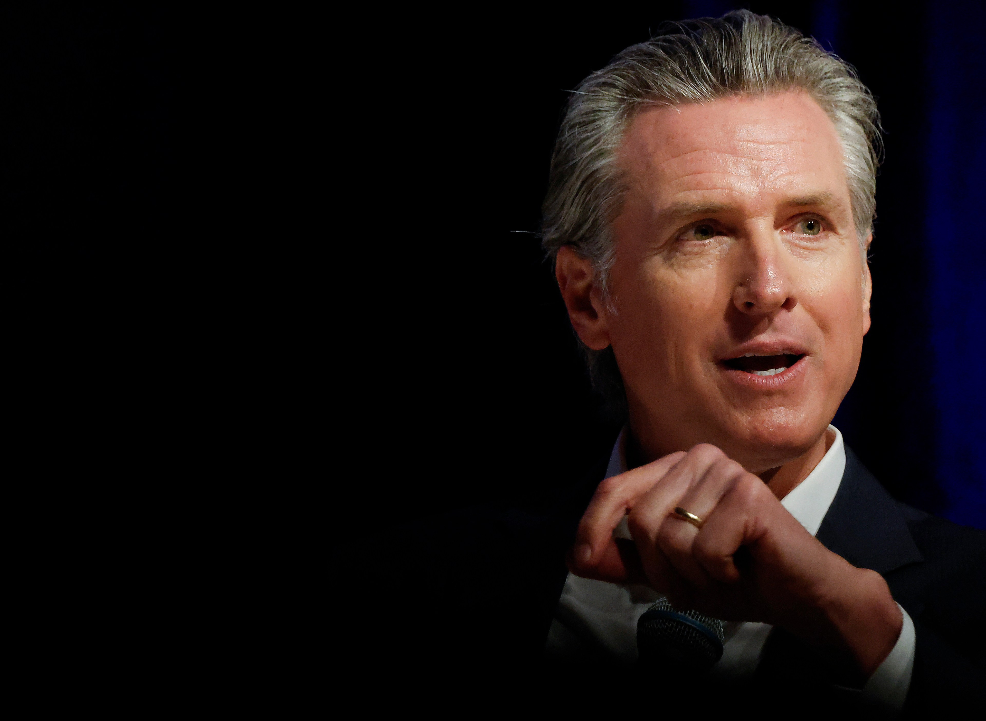 A man with gray hair is speaking in a dimly lit setting, wearing a suit and tie. Only his upper body is visible against a dark background.