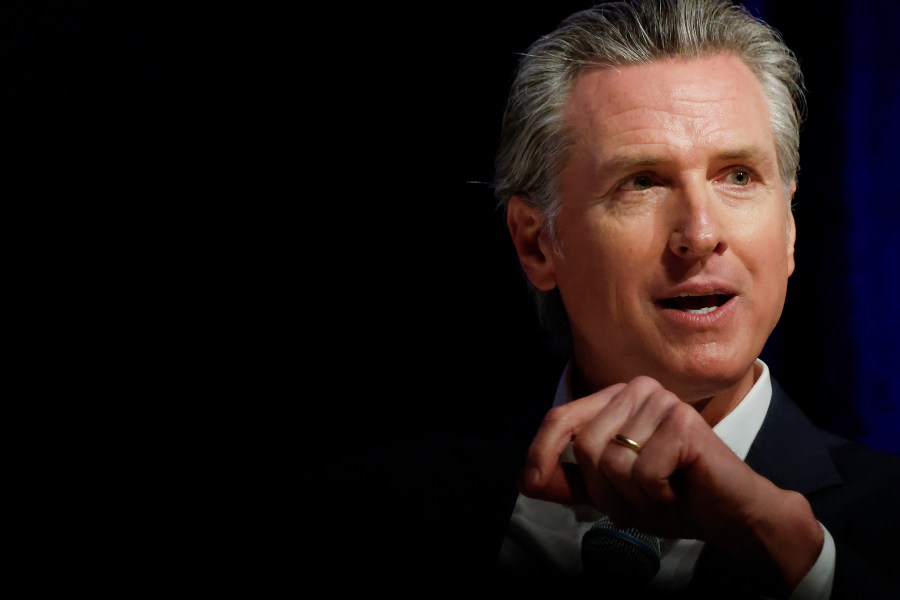 A man with gray hair is speaking in a dimly lit setting, wearing a suit and tie. Only his upper body is visible against a dark background.