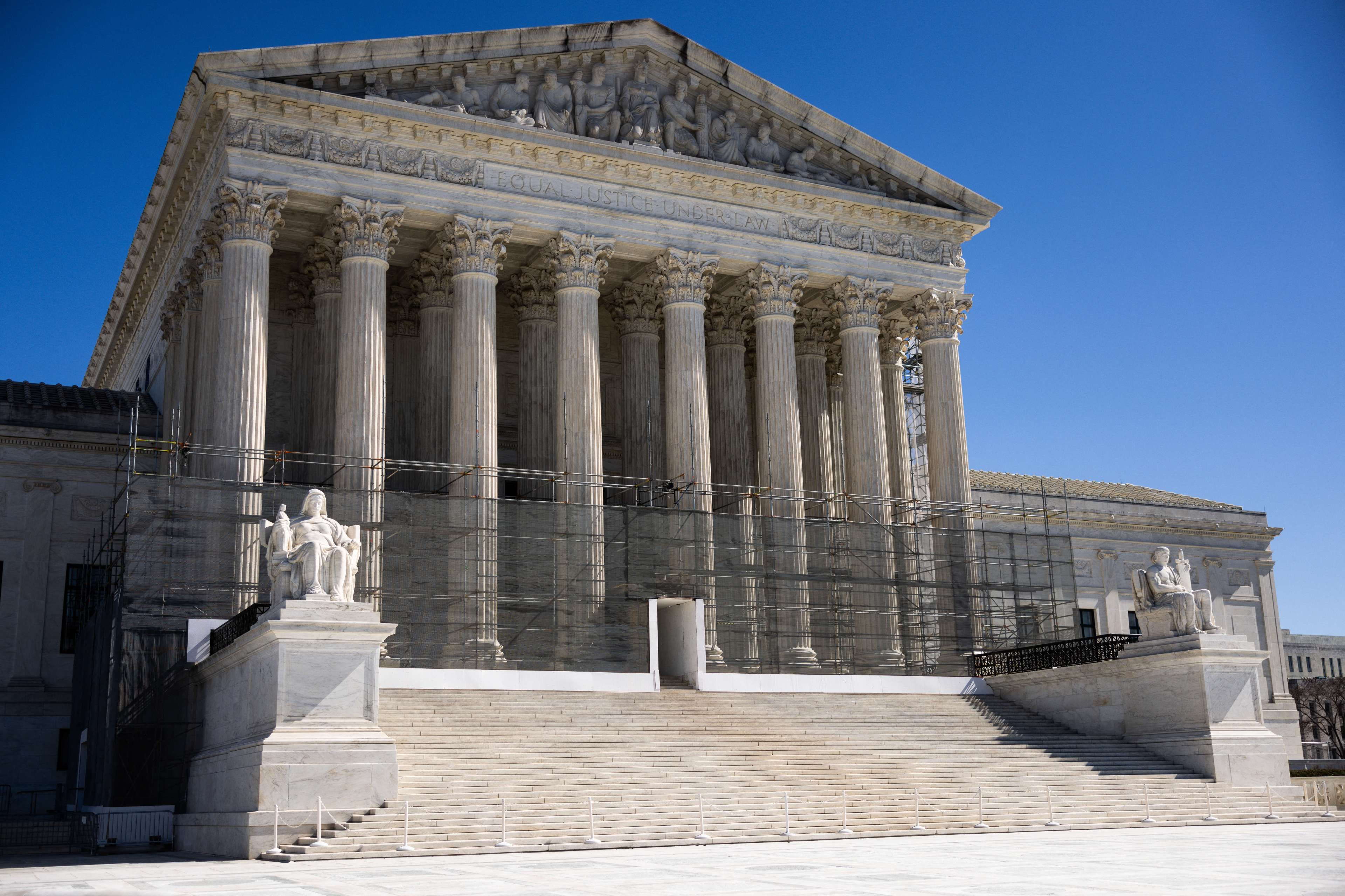 The image shows a large, classical building with grand columns and steps leading up to it. Statues flank the entrance, and a clear blue sky is in the background.