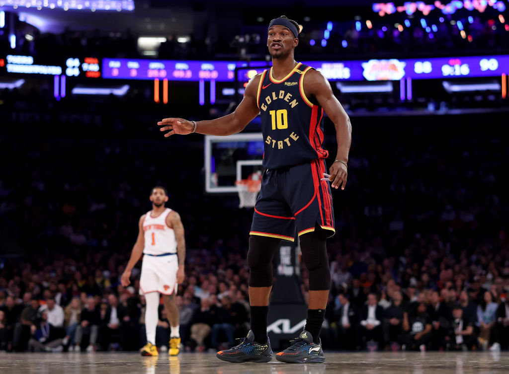 A basketball player in a &quot;Golden State&quot; jersey stands on the court. Another player in a white jersey is blurred in the background. The arena is dimly lit.