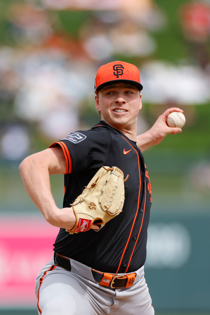 A baseball player in a black and orange uniform is pitching, wearing a matching cap and holding a glove. The background shows a blurred stadium scene.