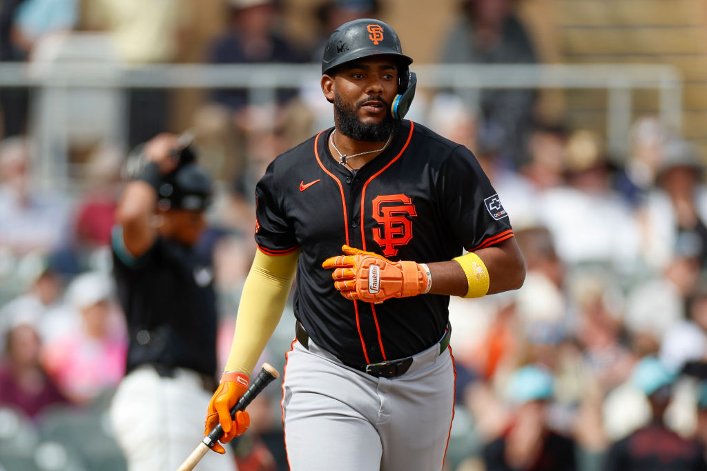 A baseball player in a black and orange Giants uniform walks on the field, holding a bat. He's wearing a helmet, gloves, and a yellow arm sleeve.