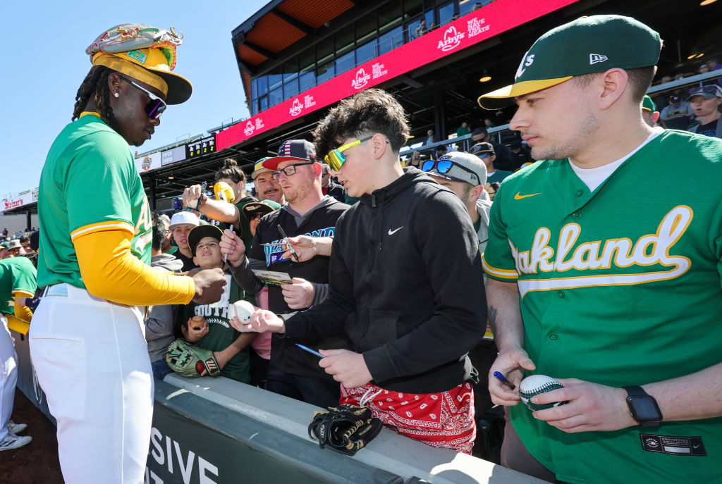 A baseball player in a green and yellow uniform signs autographs for fans near a stadium field. Fans eagerly hold out baseballs and items to be signed.