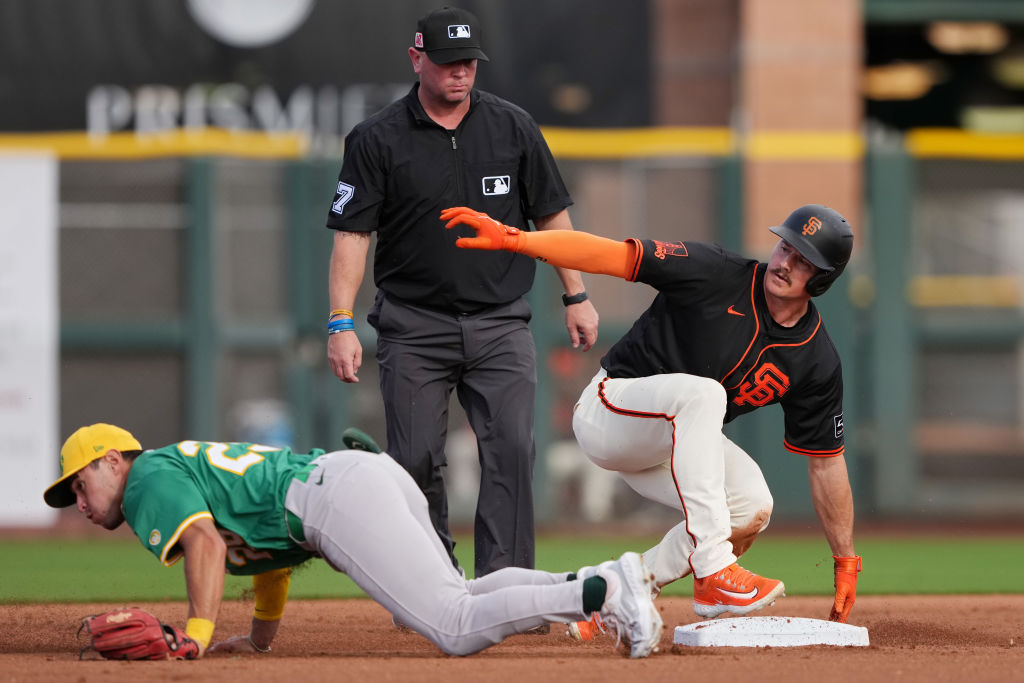 A baseball player in a black uniform slides toward a base, with another player in green attempting to tag him. An umpire in black watches closely.