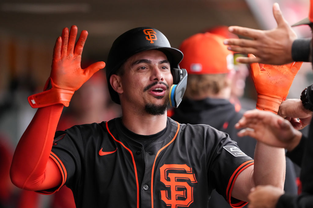 A baseball player in a black jersey and helmet, with orange gloves, celebrates high-fiving teammates in a dugout.