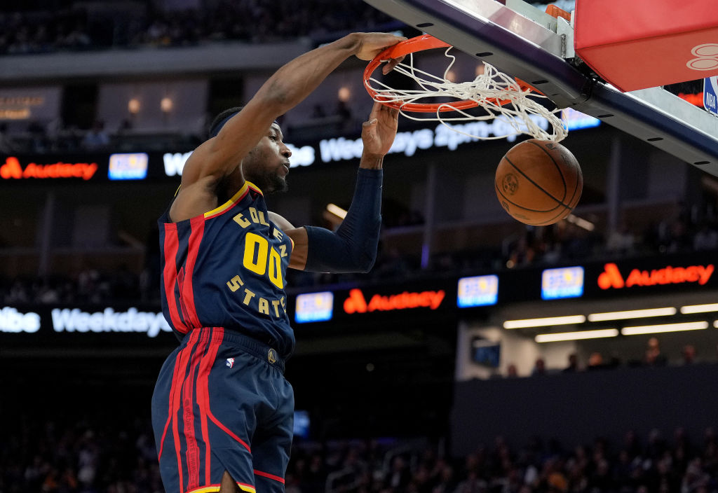 A basketball player wearing a Golden State jersey is in mid-air about to dunk the ball into the hoop. The background shows a crowded arena.