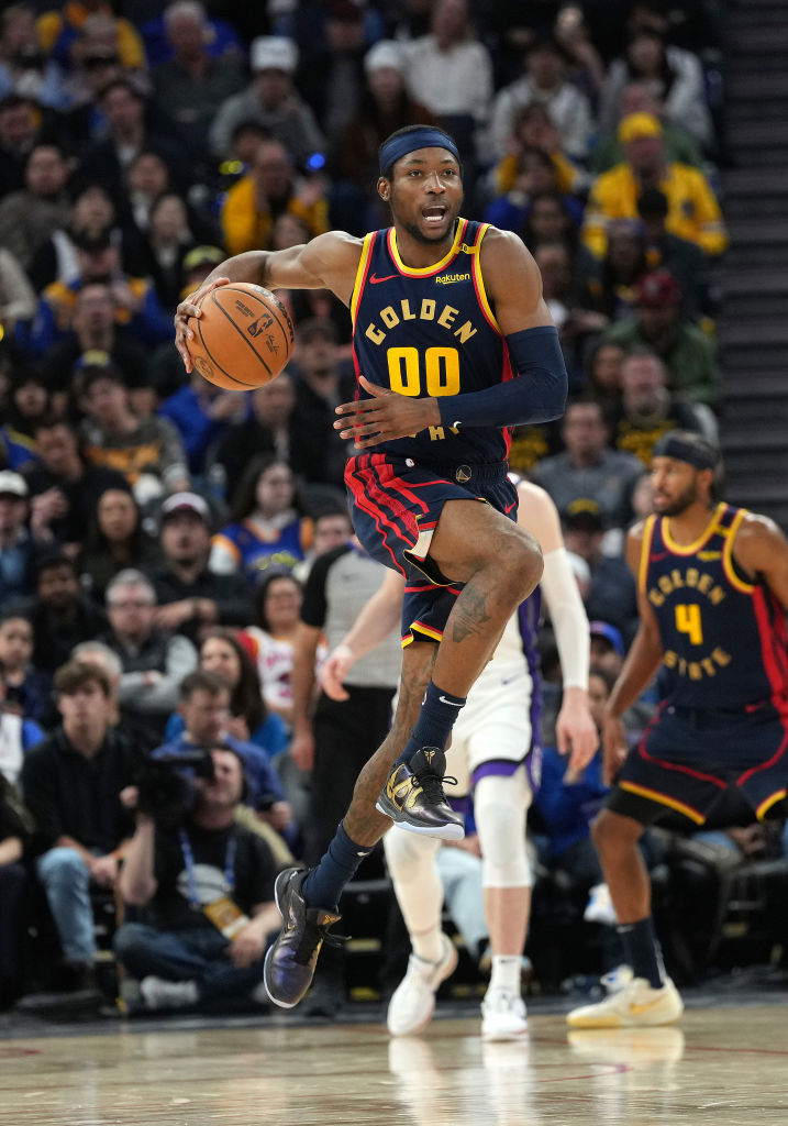 A basketball player in a &quot;Golden State&quot; jersey with number 00 is mid-air, holding a basketball. The crowd watches intently from the stands.