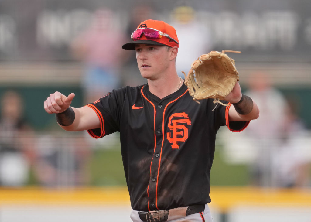 A baseball player wearing a black and orange uniform with an &quot;SF&quot; logo is on a field. He's holding a glove in one hand and gesturing with the other.