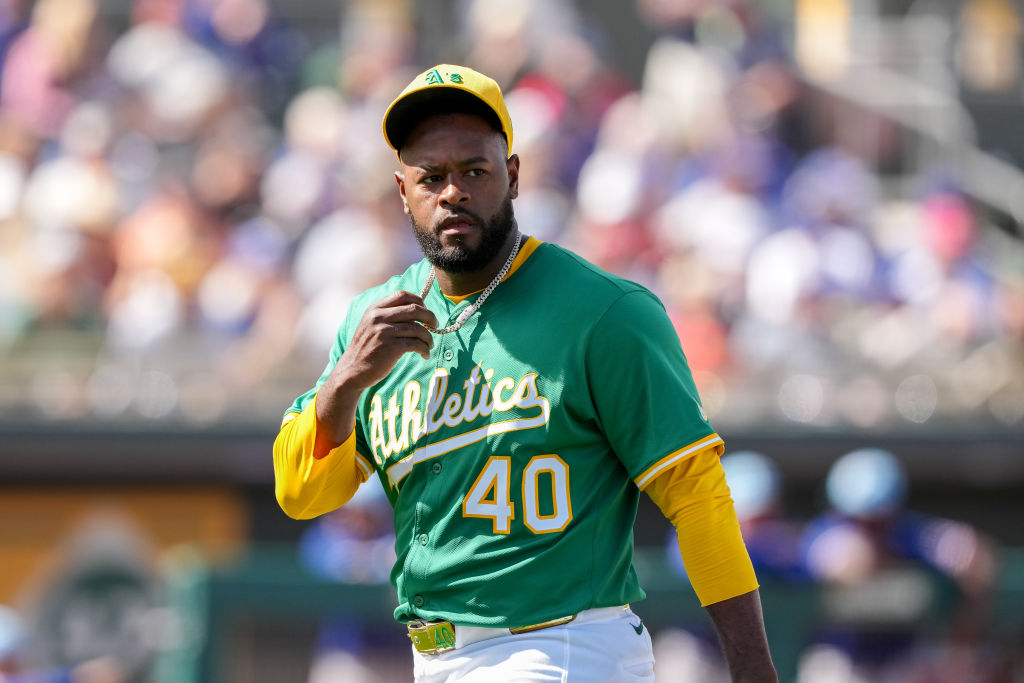 A baseball player in a green "Athletics" jersey and yellow details walks on the field, wearing a cap and number 40, with a blurred crowd in the background.