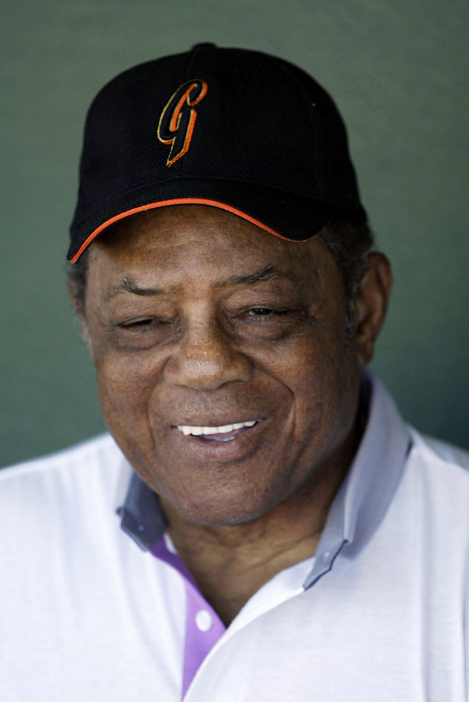 A smiling older man is wearing a black baseball cap with an orange letter and a white collared shirt with a purple accent. The background is a soft, muted green.