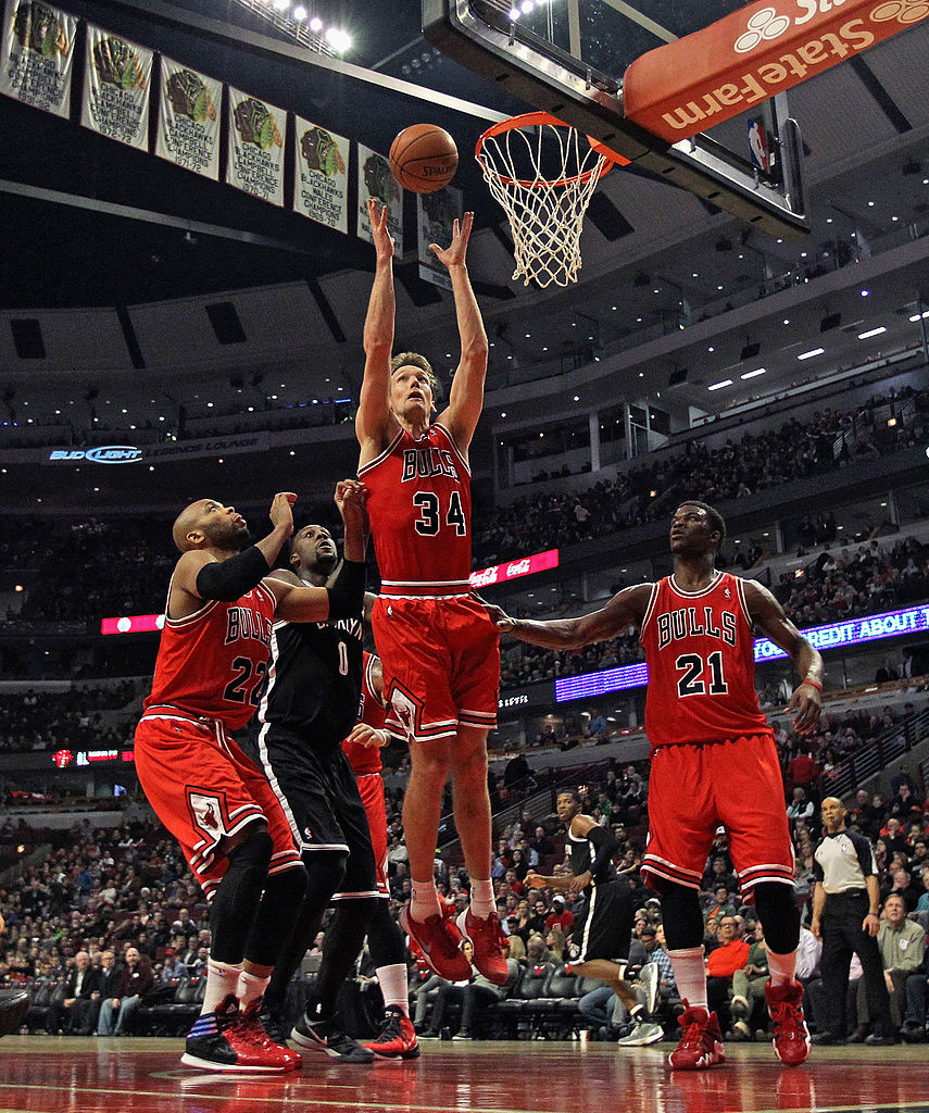 A basketball player in a red uniform jumps near the hoop, surrounded by two defenders and a teammate in a crowded arena.