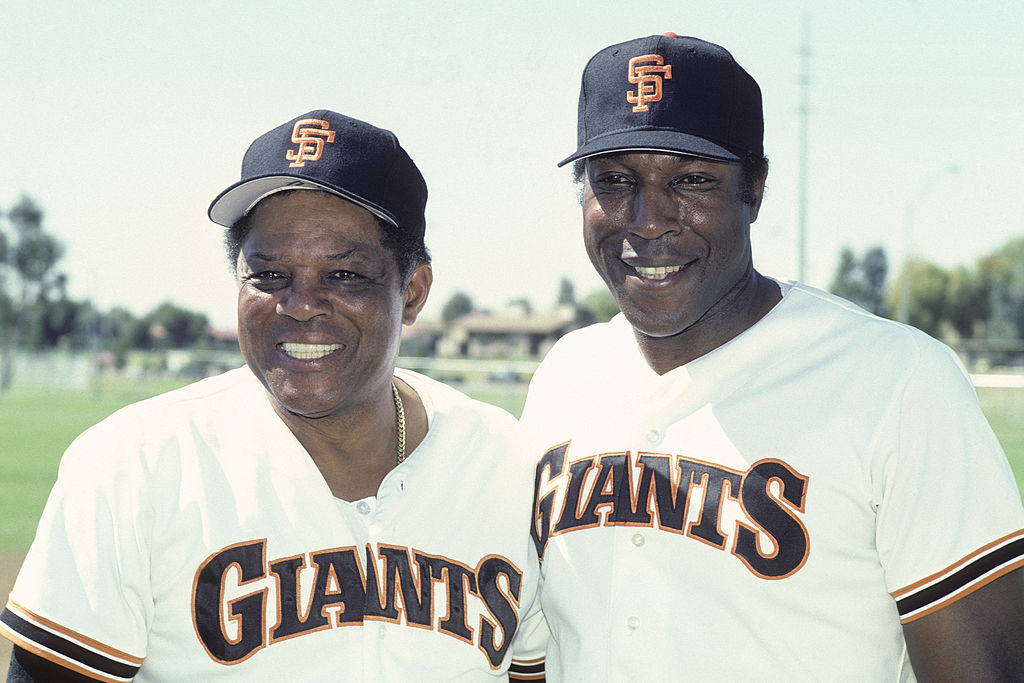 Two men wearing San Francisco Giants baseball uniforms and caps smile outdoors. The background shows a grassy field and trees.