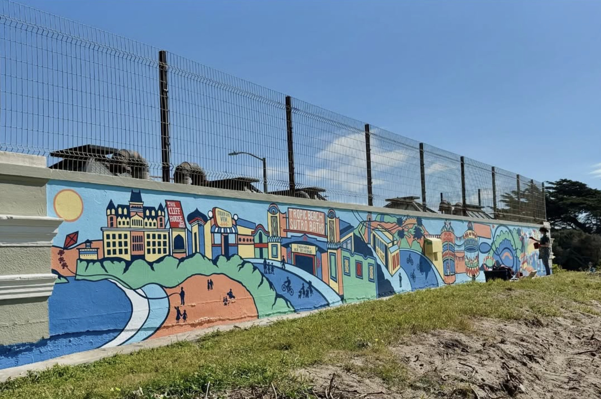 A colorful mural on a wall depicts various buildings, people, and landscapes, with signs reading &quot;The Cliff House,&quot; &quot;Sutro Baths,&quot; and &quot;Ocean Beach.&quot;