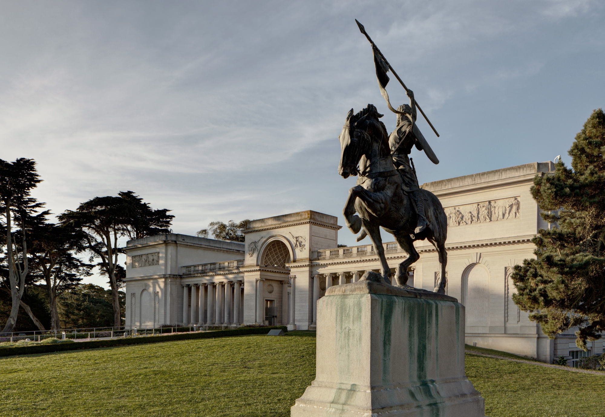 A bronze statue of a rider on a rearing horse holds a spear, positioned on a stone pedestal. Behind it is a neoclassical building with trees on the left.