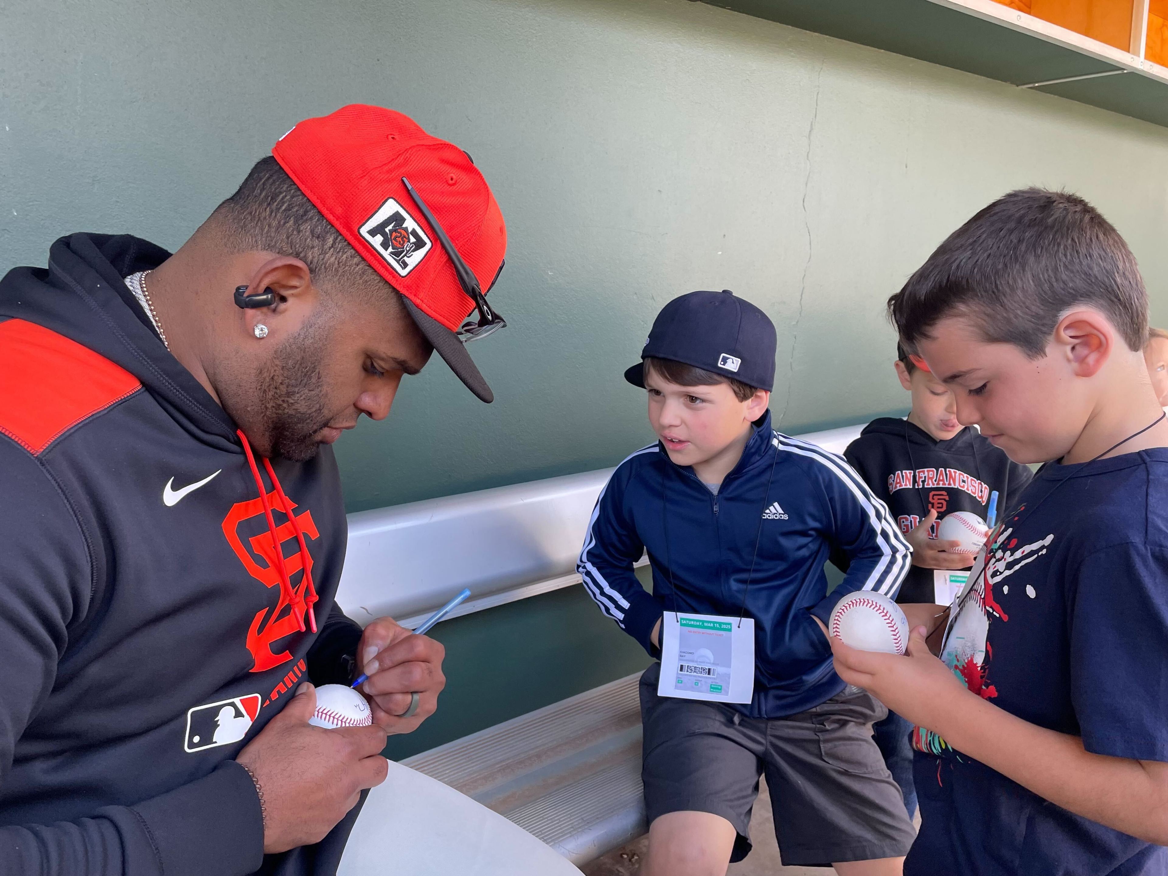 A man in a red cap is signing a baseball while two young boys watch. The boys are holding baseballs and appear excited. One wears a navy jacket and cap.