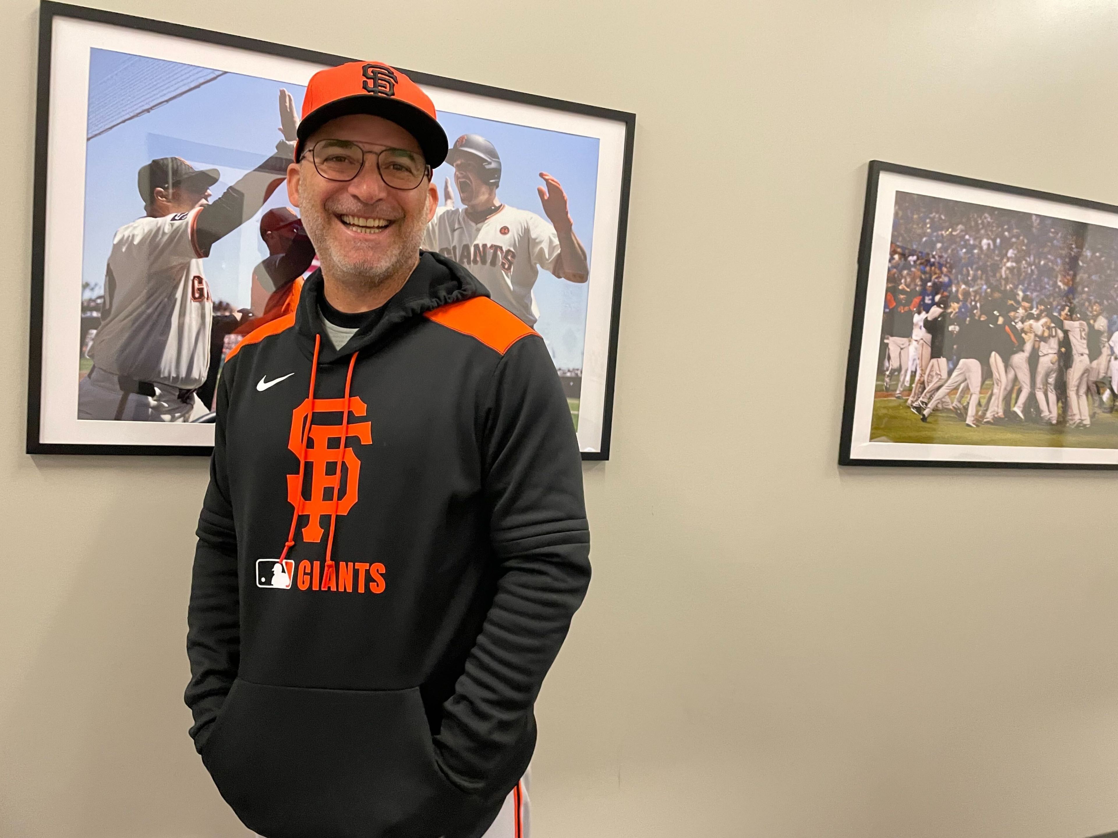 A smiling man in a San Francisco Giants hoodie and cap stands by two framed baseball photos on a beige wall.