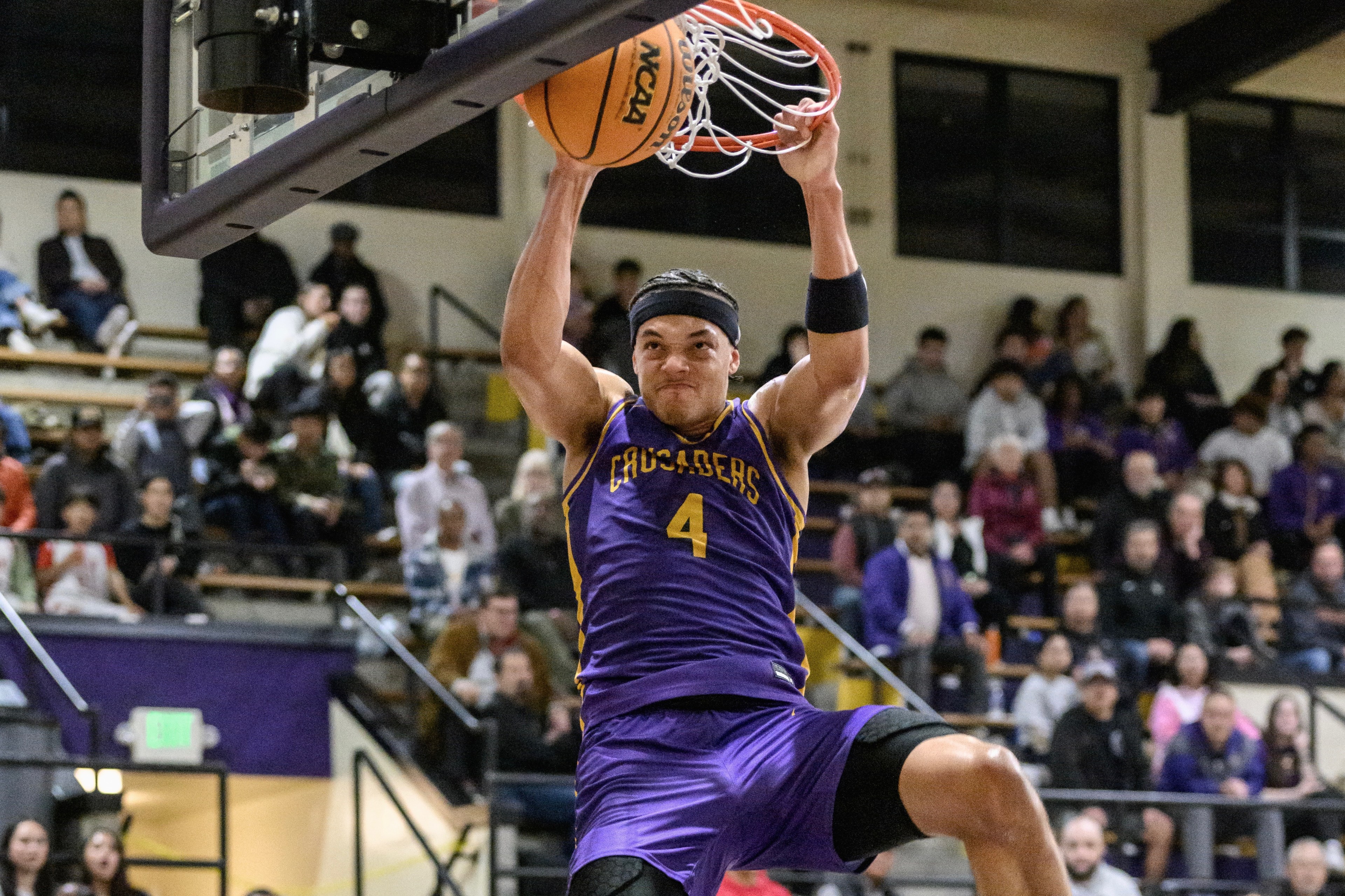 A basketball player, wearing a purple jersey with the number 4 and "CRUSADERS" written on it, is dunking the basketball during a game with a cheering crowd in the background.